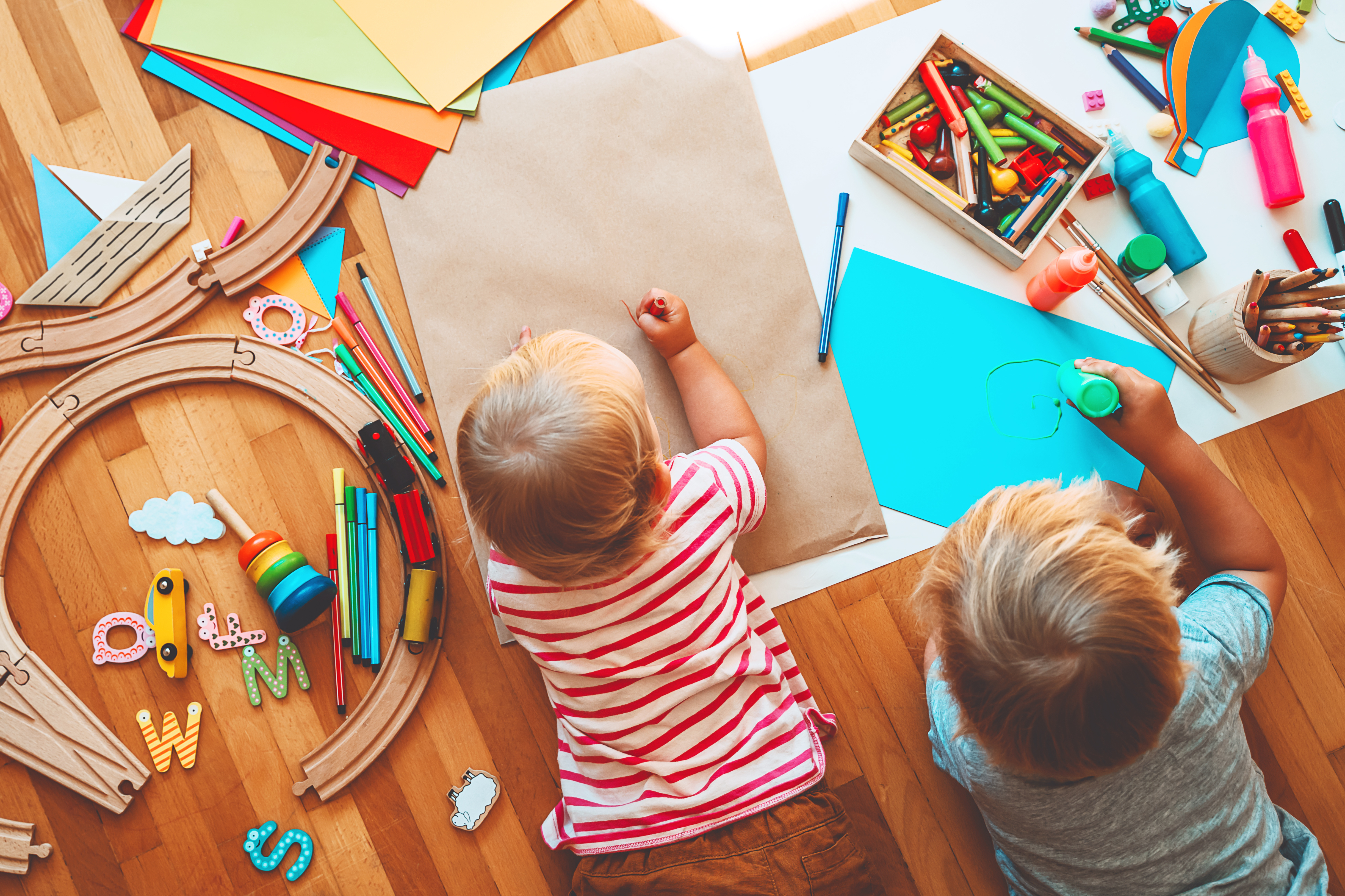 zwei Kleinkinder liegen auf dem Boden umgeben von Spielsachen und Malzubehör und malen